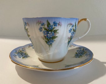 Op de afbeelding: Een witte theekop en schotelset met een lichtblauwe rand en gouden rand. De kop en schotel zijn versierd met trossen blauwe en roze bloemen. Het handvat van de kop is goudkleurig.