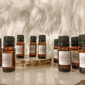 May include: A collection of 10 small brown glass bottles with black lids and white labels. The bottles are arranged on a gold-coloured decorative stand. The labels on the bottles read "Milan Scents".