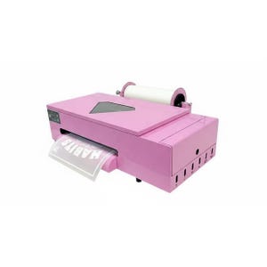 May include: A light pink desktop printer featuring a top-mounted white paper roll and a triangular viewing window. The device has a front output slot displaying a printed sheet with the text HABITS visible. The side panel includes five vertical slots.