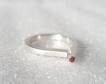 May include: A silver-toned open ring featuring a flat, rectangular band that tapers to a rounded end. A single, small, circular red gemstone is set into the tip of the rounded end, providing a distinct contrast to the metallic finish of the band.