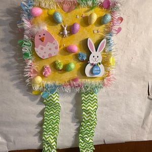Puede incluir: Adorno de pared de Pascua amarillo con varios elementos decorativos. Presenta un huevo rosa y blanco, un conejito blanco y varios huevos coloridos. Los bordes están adornados con flecos coloridos y cintas de chevron verdes y blancas.