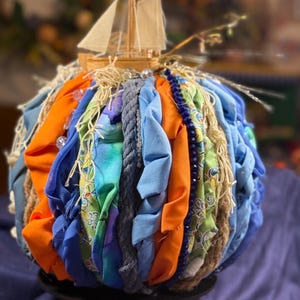 May include: A decorative sphere made from fabric strips in blue, orange, and green, with rope and beads. A small wooden sailboat is on top. The sphere sits on a black base, creating a nautical theme. The sphere's diameter is approximately 20 cm.