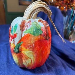 May include: A decorative pumpkin with a painted design featuring fall foliage and berries. The pumpkin is primarily orange and yellow, with a light-colored stem. The design includes green leaves and red berries, perfect for autumn decor.