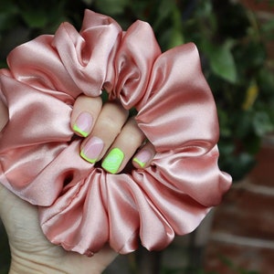 May include: A pink satin scrunchie held in a hand with neon green nail polish.