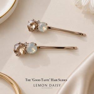 May include: Two gold-tone metal hair slides, each featuring one large champagne-coloured crystal and one smaller iridescent white crystal. The text 'The Good Taste Hair Slides' and 'Lemon Daisy Accessories' is printed below the hair clips.