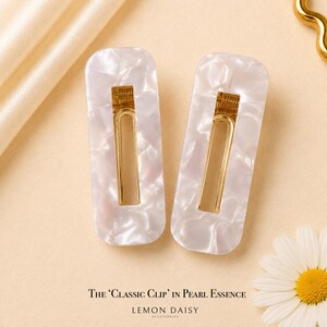 Peut inclure: Deux barrettes rectangulaires avec un design blanc nacré et doré. Les barrettes ont un effet marbré et des accents métalliques dorés. Le texte "The 'Classic Clip' in Pearl Essence" et "Lemon Daisy Accessories" est visible.