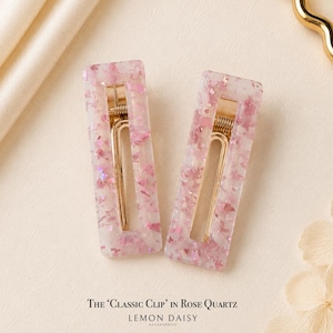 Pode incluir: Duas presilhas retangulares para cabelo com design de quartzo rosa. As presilhas têm um fecho de metal dourado e são preenchidas com glitter rosa e iridescente. O texto na parte inferior diz "THE 'CLASSIC CLIP' IN ROSE QUARTZ" e "LEMON DAISY ACCESSORIES."