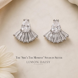 May include: A pair of silver stud earrings featuring a central pear-shaped clear stone surrounded by a fan-shaped arrangement of baguette-cut clear stones. The text below reads: The ‘She’s The Moment’ Studs in Silver, Lemon Daisy Accessories.