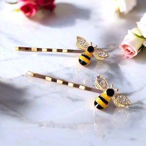May include: Two gold-coloured bobby pins, each adorned with a bee motif. The bees feature black and yellow bodies, crystal-encrusted wings, and black heads. The pins have a striped pattern and are displayed on a marble surface with pink roses.