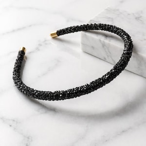 Puede incluir: Un collar tipo gargantilla negro con una superficie texturizada cubierta de pequeños cristales oscuros facetados. La banda circular está abierta en la parte delantera y rematada con tapas de metal dorado pulido en los extremos de la estructura flexible.