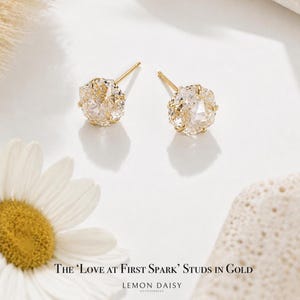 May include: A pair of gold stud earrings featuring large, clear, faceted crystals. The crystals are set in a delicate gold frame. The earrings are presented on a white surface, with a daisy and a textured stone in the background.