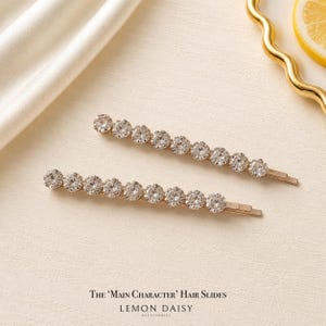 May include: Two gold-toned hair slides, each featuring a row of ten clear, round-cut faceted crystals. The hair accessories are positioned horizontally, showcasing the metallic finish and the sparkling stones. Text reads: The 'Main Character' Hair Slides, Lemon Daisy Accessories.