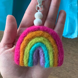 Könnte beinhalten: Ein regenbogenfarbener Filz-Schlüsselanhänger mit einem silbernen Verschluss und zwei weißen Bommeln. Der Schlüsselanhänger ist aus Filz und hat ein Regenbogen-Design mit den Farben Pink, Orange, Gelb, Grün, Blau und Lila. Der Schlüsselanhänger ist an einem silbernen Verschluss mit einer Schlaufe zum Anbringen an einer Tasche oder Schlüssel befestigt. Der Schlüsselanhänger ist handgefertigt und perfekt, um jeder Tasche oder jedem Schlüssel einen Hauch von Verspieltheit zu verleihen.