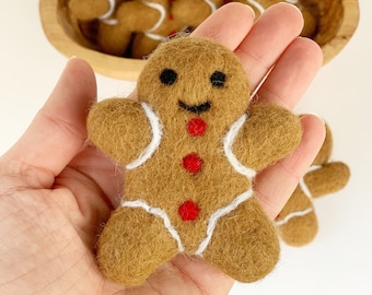 May include: A felt gingerbread man with white icing details and three red button accents is held in a hand. Several more gingerbread men are in a wooden bowl in the background. The gingerbread men have black smiles and are a festive holiday decoration.