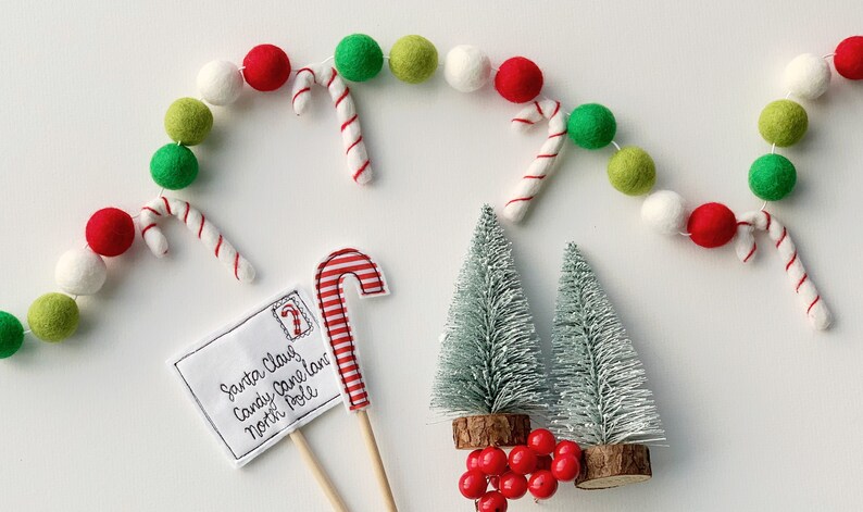 May include: A white, green, and red felt ball garland with candy canes hangs above a white envelope with the text "Santa Claus Candy Cane Lane North Pole". Two small, silver artificial Christmas trees with red berries are in the bottom right corner.
