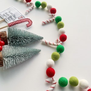 May include: A white, green, and red felt ball garland with candy cane accents. The garland is arranged in a semi-circle shape. There are two small artificial Christmas trees and a sign that says "Santa Claus Candy Cane North Pole" in the image.