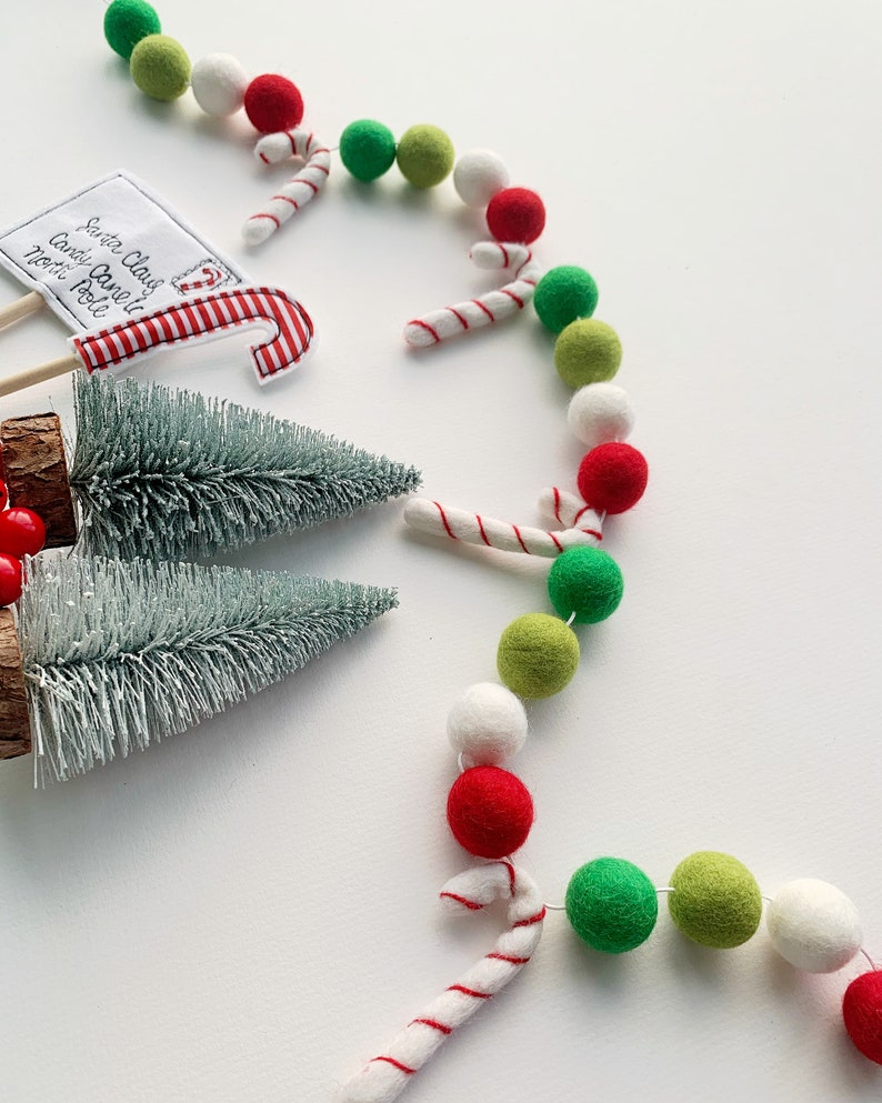 May include: A white, green, and red felt ball garland with candy cane accents. The garland is arranged in a semi-circle shape. There are two small artificial Christmas trees and a sign that says "Santa Claus Candy Cane North Pole" in the image.