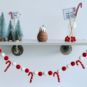 May include: A white shelf with a red and white felt ball garland hanging from it. The garland has red and white candy canes attached. There are three small artificial Christmas trees on the shelf, a small gingerbread man figurine, and a glass bottle with a red and white candy cane and a note that says "Santa Claus Candy Cane North Pole".
