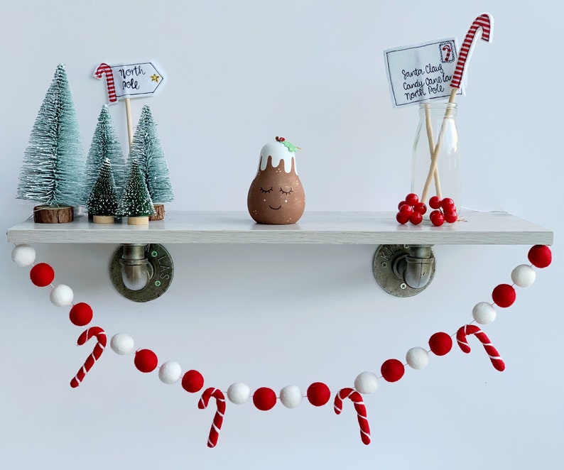 May include: A white shelf with a red and white felt ball garland hanging from it. The garland has red and white candy canes attached. There are three small artificial Christmas trees on the shelf, a small gingerbread man figurine, and a glass bottle with a red and white candy cane and a note that says "Santa Claus Candy Cane North Pole".