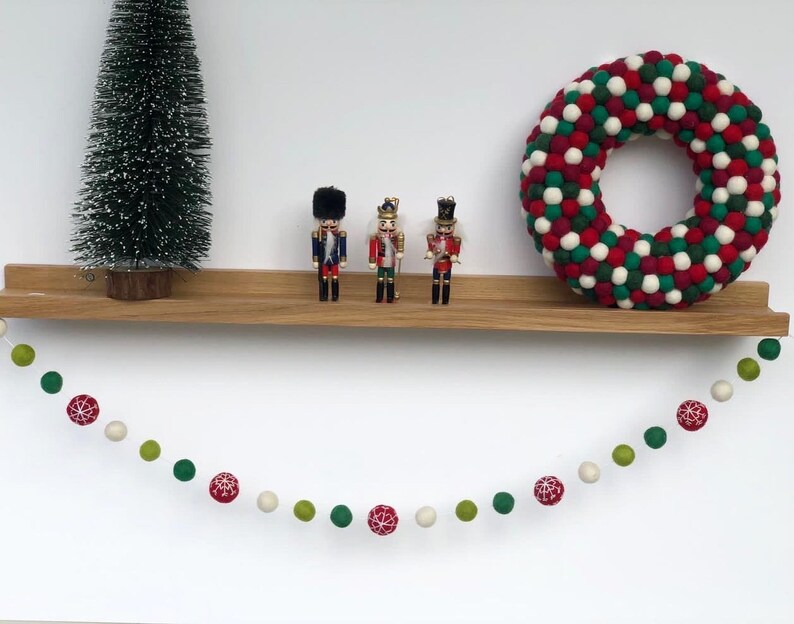 Op de afbeelding: Een guirlande van rode, groene en witte viltballen hangt aan een houten plank. Drie notenkrakerfiguurtjes staan op de plank, en een rode, groene en witte viltballenkrans hangt aan de muur achter de plank.