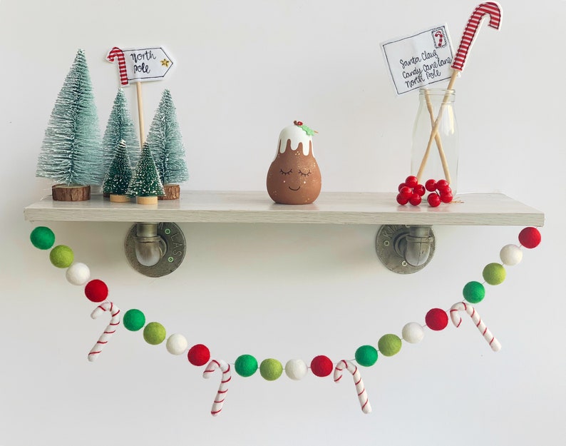 May include: A festive garland with green, white, and red felt balls and candy cane accents. The garland is hanging from a shelf with a small Christmas pudding figurine, a miniature Christmas tree, and a note addressed to Santa Claus.