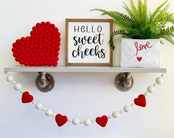Könnte beinhalten: Ein weißes Regal mit einem roten Filzherz, einem Schild mit der Aufschrift "Hello sweet cheeks" und einem weißen Umschlag mit dem aufgestickten Wort "love". Unter dem Regal hängt eine weiße und rote Girlande mit Filzherzen.