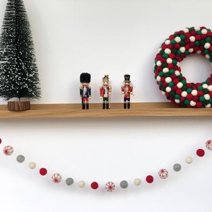 Op de afbeelding: Een houten plank met een rode en groene krans van viltballen, drie notenkrakerfiguren en een guirlande van rode en witte viltballen die aan de plank hangt.