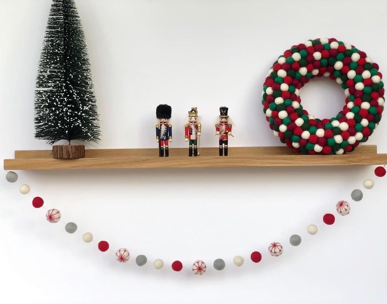 Op de afbeelding: Een houten plank met een rode en groene krans van viltballen, drie notenkrakerfiguren en een guirlande van rode en witte viltballen die aan de plank hangt.