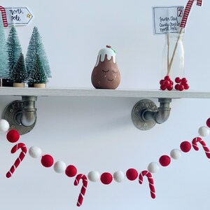 May include: A red and white felt ball garland with candy cane accents hangs from a white shelf with a small Christmas tree, a pudding figurine, and a glass jar with a candy cane and a note that reads "Santa Claus Candy Cane Lane North Pole".