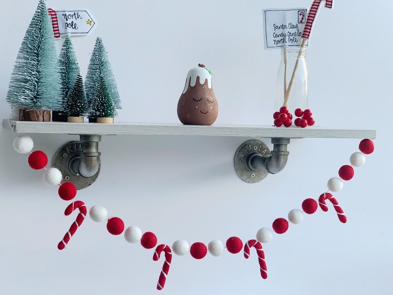 May include: A red and white felt ball garland with candy cane accents hangs from a white shelf with a small Christmas tree, a pudding figurine, and a glass jar with a candy cane and a note that reads "Santa Claus Candy Cane Lane North Pole".