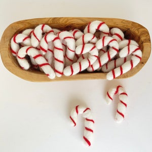 May include: A wooden bowl filled with white felt candy canes with red stripes. The candy canes are arranged in a random pattern. Two additional candy canes are on a white surface.