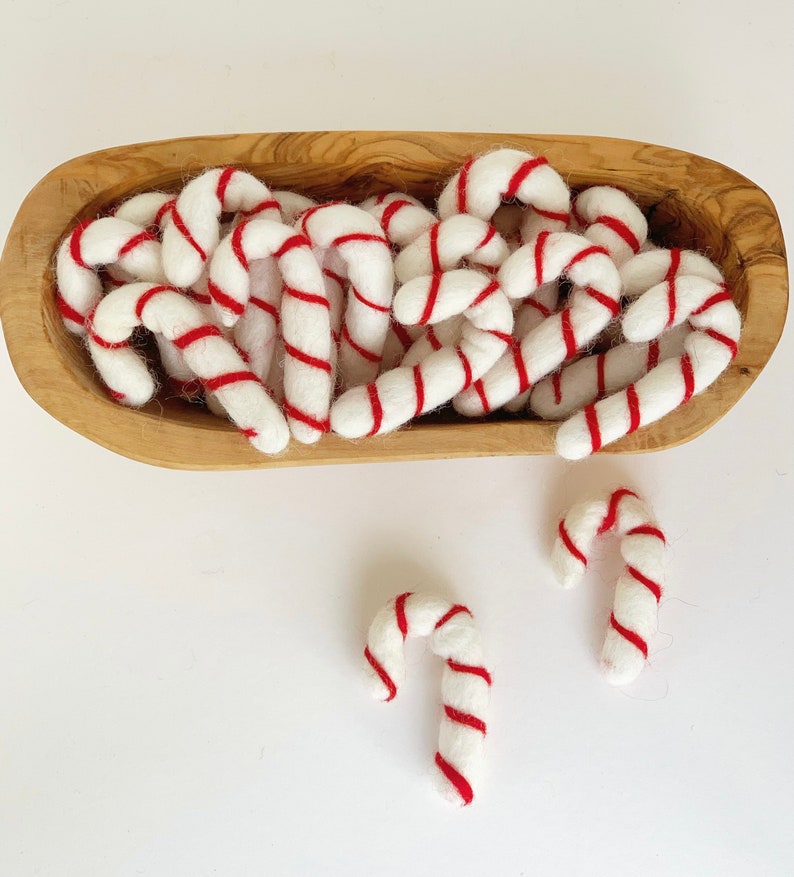 May include: A wooden bowl filled with white felt candy canes with red stripes. The candy canes are arranged in a random pattern. Two additional candy canes are on a white surface.