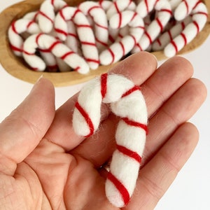 May include: A close-up of a hand holding a white and red felt candy cane ornament. The candy cane is made of wool felt and has a red and white striped pattern.
