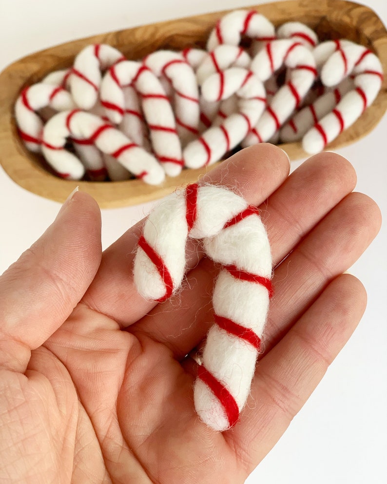 May include: A close-up of a hand holding a white and red felt candy cane ornament. The candy cane is made of wool felt and has a red and white striped pattern.