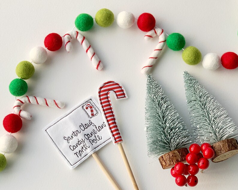 May include: A white envelope with the text "Santa Claus Candy Cane Lane North Pole" and a red and white candy cane on a stick. There are two small green trees with red berries and a garland of white, red, and green felt balls with candy canes.