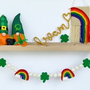 May include: A wooden shelf with two gnomes, a rainbow wall hanging, and a felt rainbow garland with shamrock accents. The garland reads "dream".