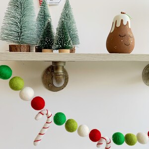 May include: A festive garland made of green, red, and white felt balls with candy cane accents. The garland is hanging on a white wall with a shelf above it. The shelf has a small Christmas tree, a sign that says "North Pole", and a small brown and white Christmas pudding ornament.