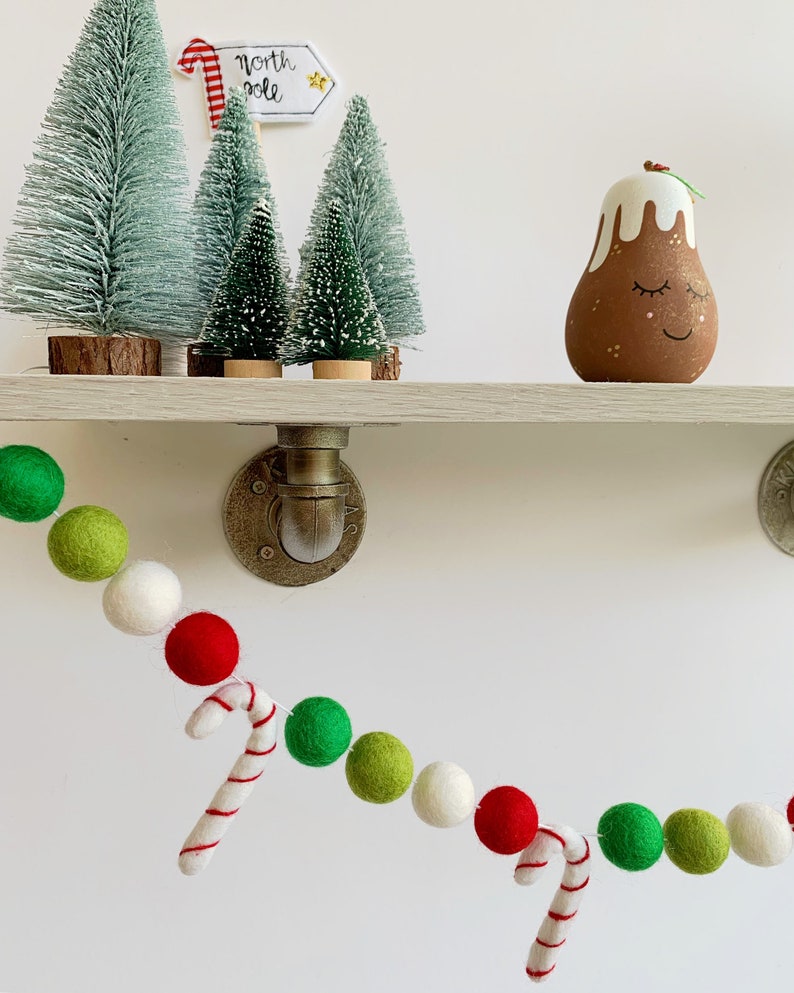 May include: A festive garland made of green, red, and white felt balls with candy cane accents. The garland is hanging on a white wall with a shelf above it. The shelf has a small Christmas tree, a sign that says "North Pole", and a small brown and white Christmas pudding ornament.