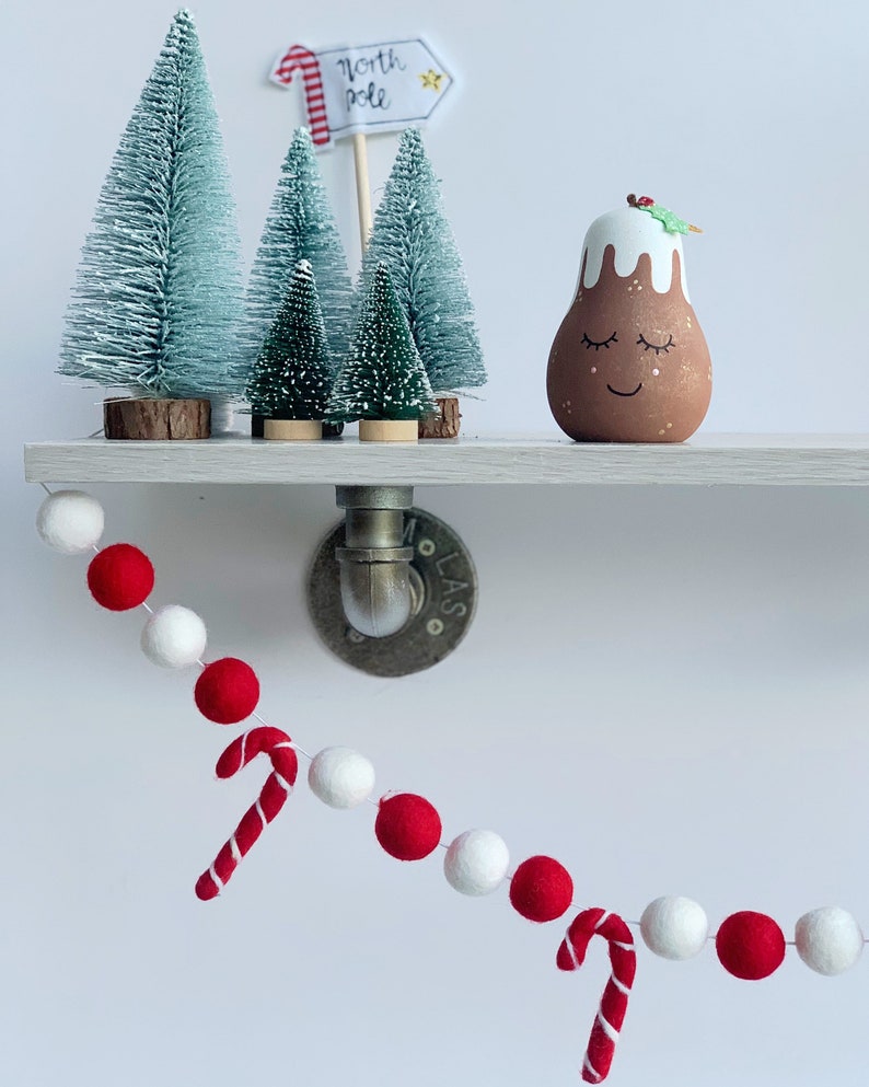 May include: A white shelf with a red and white felt ball garland with candy cane accents hanging from it. There are three small green artificial Christmas trees and a brown and white Christmas pudding ornament on the shelf. The sign above the shelf reads "North Pole".