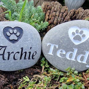 Op de afbeelding: Twee grijze stenen met gegraveerde poot afdrukken in harten en de namen "Archie" en "Teddy".