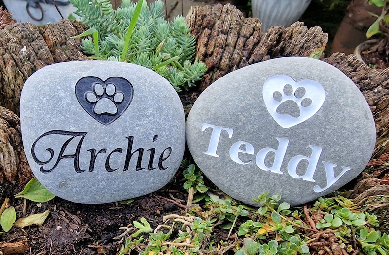 Op de afbeelding: Twee grijze stenen met gegraveerde poot afdrukken in harten en de namen "Archie" en "Teddy".