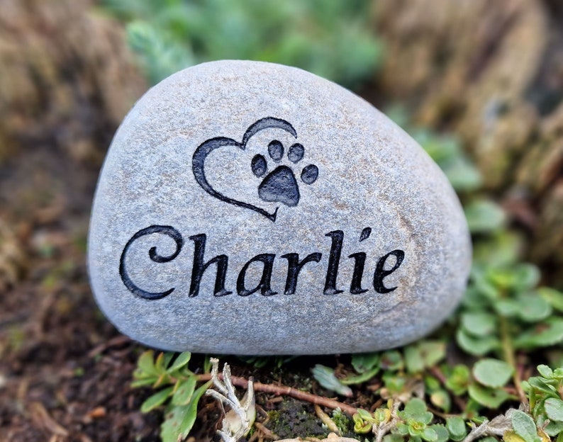 Op de afbeelding: Een grijze steen gegraveerd met een hart en een pootprint, en de naam "Charlie".