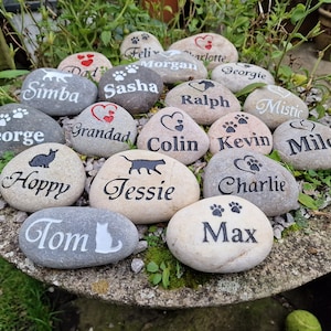 Op de afbeelding: Een verzameling van 16 gepersonaliseerde huisdier-gedenkstenen met verschillende namen en poot afdrukken. De stenen zijn grijs en beige met zwarte en rode letters.