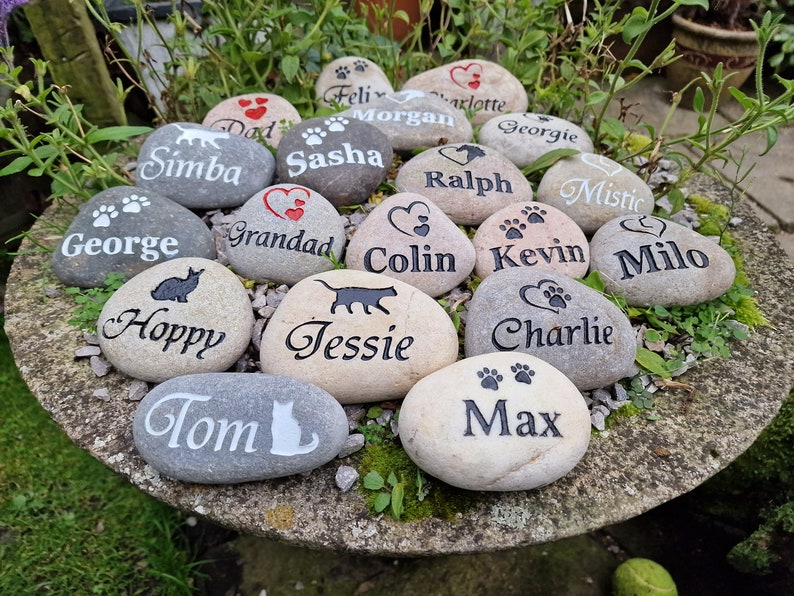 Op de afbeelding: Een verzameling van 16 gepersonaliseerde huisdier-gedenkstenen met verschillende namen en poot afdrukken. De stenen zijn grijs en beige met zwarte en rode letters.
