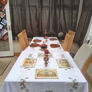 May include: A white tablecloth with embroidered Christmas designs covers a wooden table set for a festive meal. The table is set with wooden chargers, placemats, and decorative centerpieces. Wooden chairs surround the table, and brown curtains are in the background.