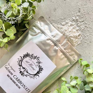May include: A silver foil pouch containing white powder labeled Amy Howard at Home Pure Organic Calcium Carbonate Made From Ground Limestone. A small pile of the white powder sits outside the pouch on a gray surface near green foliage.