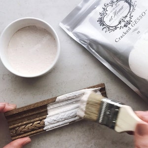 May include: A white bowl filled with a white, creamy substance. A silver bag with the text "Amy Howard at Home" and "Cracked Gesso" is in the background. A hand is applying the white substance to a wooden frame with a brush.