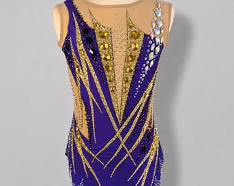 May include: A sleeveless leotard featuring a deep purple fabric base with a nude mesh neckline and upper chest panel. The garment is decorated with gold and dark purple geometric appliques, clear crystal accents, and large iridescent rhinestones.