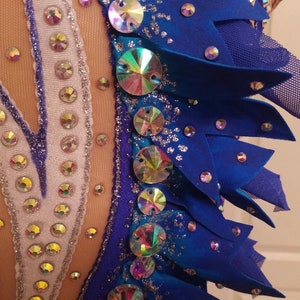 May include: Close-up of a blue leotard with silver and iridescent rhinestones. The leotard has a V-shaped neckline and a blue and white design. The fabric is sparkly and has a textured look.