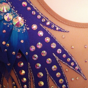 May include: A close-up of a purple leotard with a blue and teal feather design. The leotard is decorated with iridescent rhinestones.